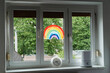 © nataliaderiabina - A bright window with a colorful painted rainbow on the glass, a clock, and a small humidifier placed on the windowsill, with green trees visible outside.