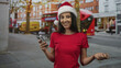 © Krakenimages.com - Woman holding smartphone and smiling on city street in red santa hat and red shirt; holiday cheer warmth.