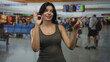 © Krakenimages.com - Young hispanic woman in casual tank top shows ok sign with her hand and points finger toward camera in busy airport terminal; confidence.