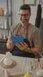 © Krakenimages.com - Man taps blue tablet at pottery table in studio wearing apron and glasses while seated among tools and ceramics; calm contentment.