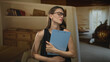 © Krakenimages.com - Woman clutching a blue clipboard to her chest while wearing glasses inside a building living room, eyes closed with a pained frown and crossed arms; concern reflection.