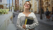 © Krakenimages.com - Blond woman with glasses holds books and a black notebook to her chest smiling with head tilt on a sunny city street lined with buildings and lampposts; joy study confidence.