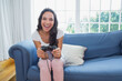© wavebreak3 - Female sitting on blue sofa in living room in white top pink pants holding wired controller