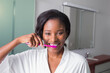 © wavebreak3 - African American woman brushing teeth at vanity with pink toothbrush, wearing white bathrobe