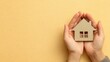 © MKS - A small wooden house model is being gently held by two hands in front of a beige background