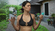 © Krakenimages.com - Woman in a black bikini top shrugs holding deodorant stick by building entrance outdoors; uncertainty.