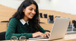 © PRASANNAPIX - A young Indian woman in a green sweater is smiling as she types on her laptop in an empty lecture hall