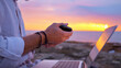© stockcopter - A digital nomad works on a laptop at sunset, holding a portable device. The serene ocean backdrop and rocky terrain highlight remote work freedom, blending technology with nature's tranquility.