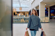 © we.bond.creations - Young woman smiling while walking through a bright modern shopping mall, carrying paper shopping bags after purchasing