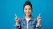 © Artomoro - Cheerful woman in denim jacket pointing upwards on blue background, perfect for announcements, promotions, or highlighting key information today