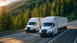 © Maksym - Perspective from asphalt level showing two white semi-trucks advancing down a highway, sun reflecting off the road surface, green forest stretching along both sides, emphasizing mo