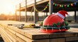 © Nina - Festive red construction hard hat adorned with a Santa hat, holly, and string lights, resting on wooden planks at a building site during golden hour.