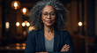 © Miftakhul - Confident African American businesswoman with grey curly hair and glasses, wearing a blazer, stands with arms crossed