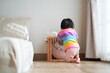 © Siriluk - A young child wearing a colorful striped shirt kneels on the wooden floor in a cozy bedroom, looking toward a small wooden crib with soft toys.