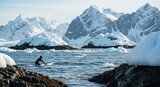 An adventurous surfer in a black wetsuit paddles through frigid blue water with icebergs, against a stunning panoramic backdrop of a vast, snowy mountain range