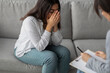 © Prostock-studio - A woman sits on a sofa, crying and grieving while discussing her feelings with a psychologist. She shows signs of stress and depression in a mental health clinic setting.