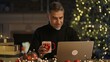 © nyul - Happy middle aged man with laptop computer at home, sitting at desk in Christmas decorated cosy room. Fireplace, Christmas tree in background. Buying gifts in web shop, searching presents online.