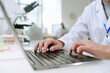© AnnaStills - Caucasian man working on laptop in laboratory, hands typing on keyboard with microscope and scientific equipment visible in background, medical research setting