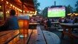 © Pete - Glass of beer on wood table at outdoor pub during soccer game. People watch sport on screen. Party outside on summer with friends cheers together.