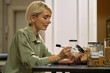 © DragonImages - Caucasian middle aged woman sitting at table labeling food storage containers with marker, organizing pantry items, focusing on task, short blond hair visible, kitchen background