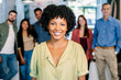 © Xavier Lorenzo - Confident young African American woman smiling in office with diverse team in background. Leader standing proudly in a modern workspace. Positivity and professional empowerment concept.