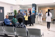 © DC Studio - Busy hospital lobby with patients and visitors seated in waiting area. Receptionist and nurse assist at front desk as diverse individuals prepare for medical appointments and consultations.