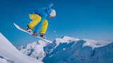 CLOSE UP, LOW ANGLE VIEW: Snowboarder launches off a steep ridge and flies above scenic snowy mountains. Clear blue sky, rugged alpine peaks and flying powder snow make this a classic freeride moment.