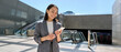 © insta_photos - Young happy Asian business woman wearing suit holding mobile phone standing in city subway using smartphone for texting, checking apps for public transport, metro or travel guide