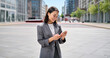 © insta_photos - Young happy Asian business woman wearing suit holding mobile phone standing in big city on busy street, smiling lady worker using smartphone for texting, cellphone apps for work outside, vertical