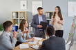 © Studio Romantic - Smiling man and woman standing near whiteboard and explaining ideas to colleagues during team meeting. Group of business professionals listening carefully and taking part in brainstorming session.