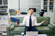 © Nuttapong punna - Asian businessman sitting on office sofa using smartphone and laptop, smiling while working with documents in a modern workspace.