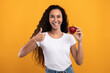 © Prostock-studio - A happy millennial woman with curly hair stands against a yellow-orange background. She holds a red apple in one hand and gives a thumbs up with the other. She promotes healthy eating.