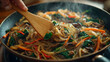 © inayah - A realistic close-up of Korean japchae (glass noodles stir-fried with vegetables and beef) cooking in a frying pan. A wooden spatula is held by the hand of a 60-year-old Korean housewife, her hand an