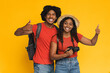 © Prostock-studio - Two smiling individuals stand together with thumbs up, enjoying a playful moment in front of a vibrant yellow backdrop. They are dressed casually, ready for an adventure with a camera in hand.