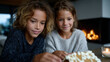 © Milos - Two children are intently decorating a gingerbread house, showcasing their creativity and joy during the festive season, while a cozy fireplace burns in the background.