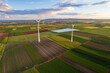 © AmazingAerialAgency - Aerial view of wind turbines standing tall amidst a patchwork of vibrant green and gold fields, contrasting with the soft hues of the sunset sky, Mannheim, Baden-Wurttemberg, Germany.