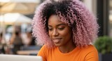 Young Black woman with pink curly hair using a laptop at an outdoor cafe. Smiling African American freelancer working remotely on a computer