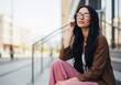 © Vasiliy - Young woman with glasses sitting on urban steps. Stylish female with long black hair in brown jacket and pink trousers outdoors. Copy space