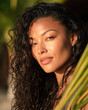 © Kowit - A close-up portrait of a woman with curly hair, glowing skin, and a serene expression, framed by green foliage in a warm, sunlit setting.