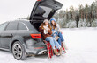 © maxbelchenko - Happy female friends sit on a blanket in the trunk of a car, having fun on a winter day. Two young women enjoy a road trip in a snowy forest. Concepts of friendship, relaxation, and adventure.