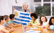 © JackF - Mature female teacher, conducting a lesson in the classroom, tells the pupils the history of Uruguay and holds the national .flag of the country in her hands