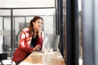 © NAMPIX - A smiling Asian woman in a red blazer sits at a table with a laptop, looking thoughtfully away with her hand on her chin, nearby window.