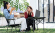 © NAMPIX - Two Asian women sit outdoors on camping chairs, laughing and gesturing animatedly during a conversation with a laptop on a table