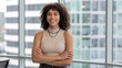 © Nimra - Smiling woman with curly hair and crossed arms standing by a window in an office setting indoors