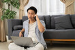 © Leny Studio - Young man sitting on floor using laptop and talking on phone in living room with sofa and curtains