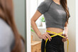© Pormezz - Weight lose, loss concept. Slim body asian young woman hand use tape measuring around waistline in fit sports. Girl looking reflect in mirror at home. Healthy nutrition, fitness for wellbeing beauty.