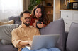 © Stockphotodirectors - Two people relax on a sofa, one using a laptop while the other holds a mug and watches with interest. The cozy living room has soft lighting and plants.