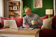 © Stockphotodirectors - A man sits comfortably on a red sofa, sipping coffee and reviewing financial documents. A laptop sits open in front of him, and the room is decorated with plants and books.
