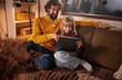© Stockphotodirectors - A father and daughter sit together on a couch, sharing laughter while looking at a tablet. The warm glow of the living room adds to the cozy winter evening atmosphere.
