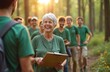 © miss irine - Happy elderly woman leads group of volunteers in forest. Diverse people wear green shirts, work on nature project. Sunny day, collaboration, teamwork.
